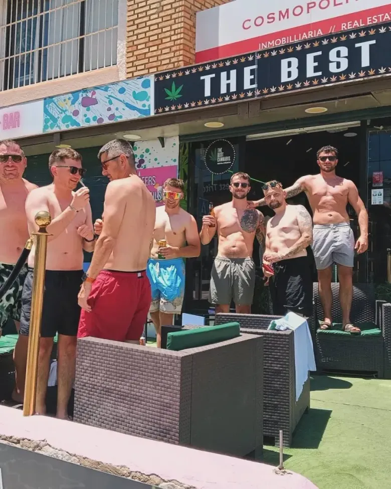 Grupo de hombres sin camisa afuera de un bar, algunos con bebidas. El cartel del bar dice 