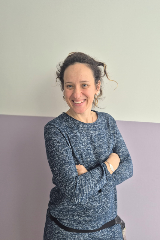 Una mujer que lleva una camiseta negra y una chaqueta gris sonríe a la cámara.