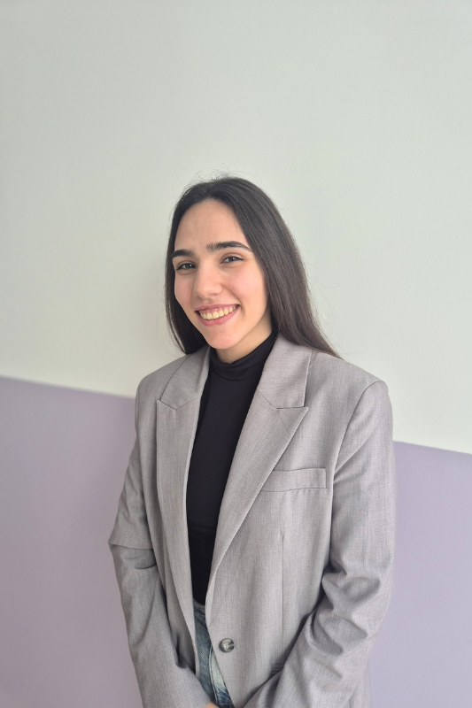 Una mujer que lleva una camiseta negra y una chaqueta gris sonríe a la cámara.