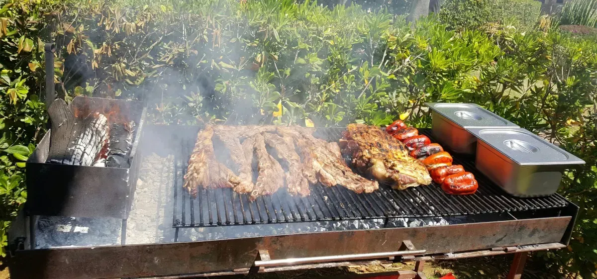 Parrilla al aire libre con varios trozos de carne y salchichas cocinándose al humo en una barbacoa grande.