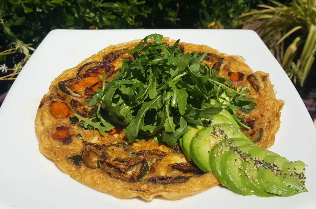 Tarta salada cubierta con verduras y rodajas de aguacate en un plato blanco al aire libre.