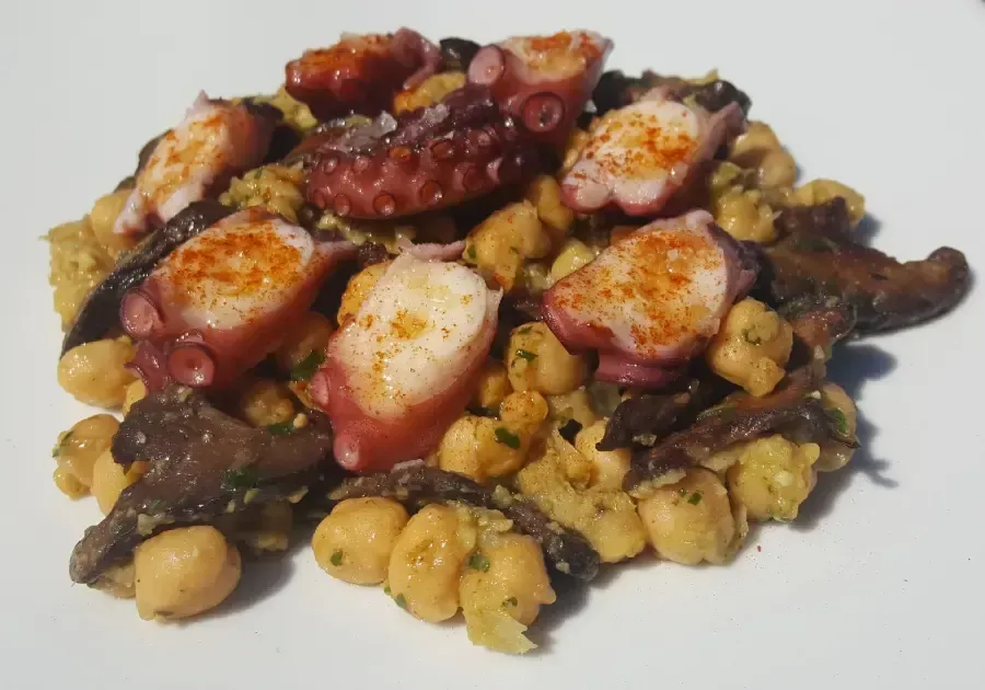 Pulpo a la parrilla con patatas y garbanzos en un plato blanco, cubierto con pimentón y aceite.