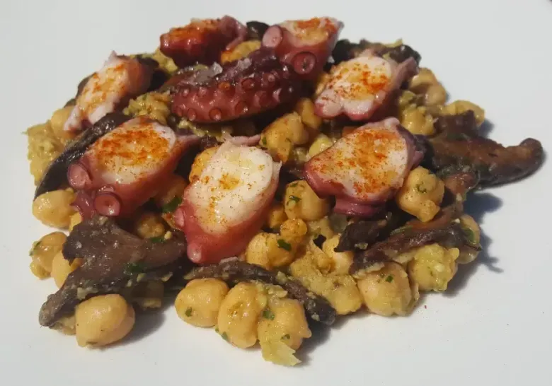 Pulpo a la parrilla sobre una cama de garbanzos en un plato blanco, rociado con salsa y condimentos.