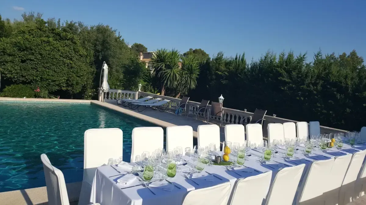 Mesa de comedor larga al aire libre junto a la piscina, con sillas blancas y vajilla azul, al lado de un muelle.