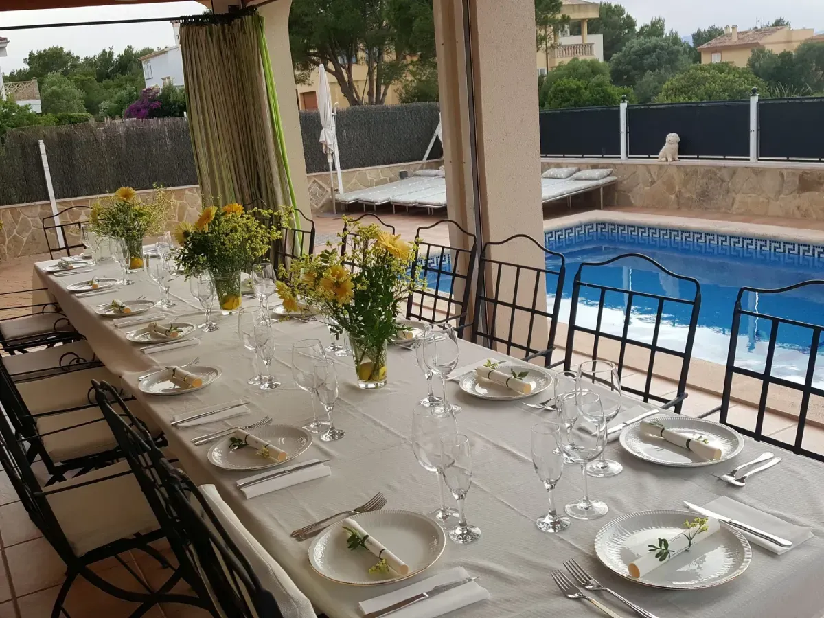 Conjunto de mesa y sillas largas para comer al aire libre con platos blancos y flores amarillas junto a una piscina.