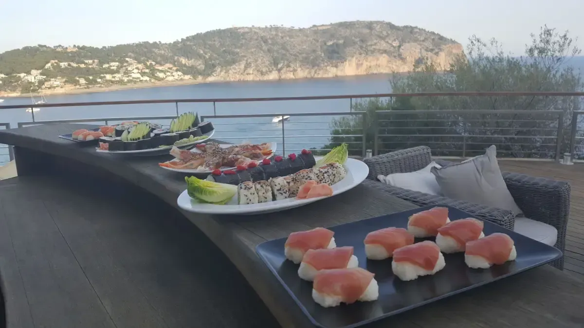 Platos de sushi en una mesa de patio con vistas a un lago tranquilo y colinas al atardecer.