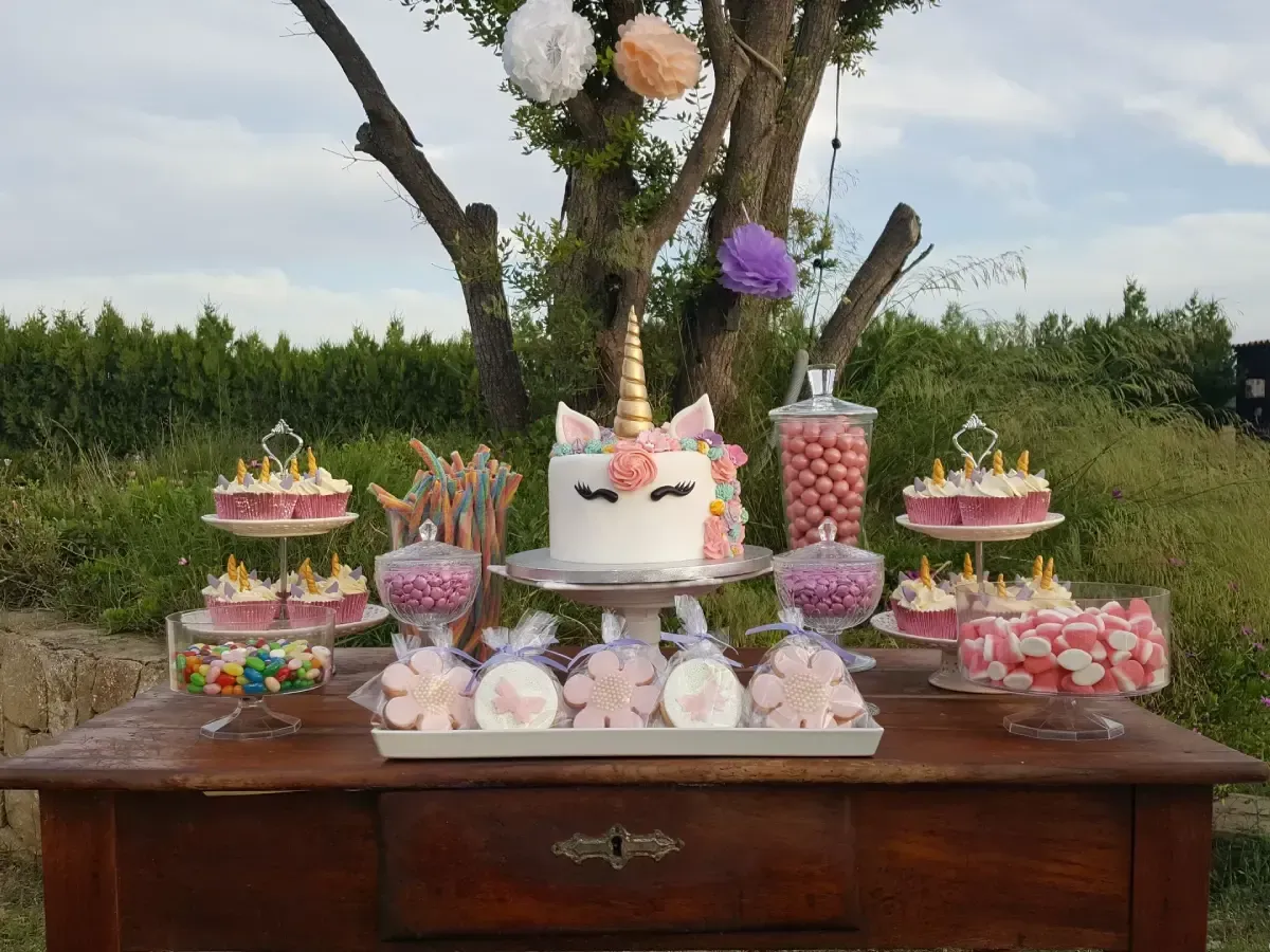 Mesa de dulces y mesa de pasteles en tonos pastel al aire libre con flores, frascos de golosinas y postres decorativos.