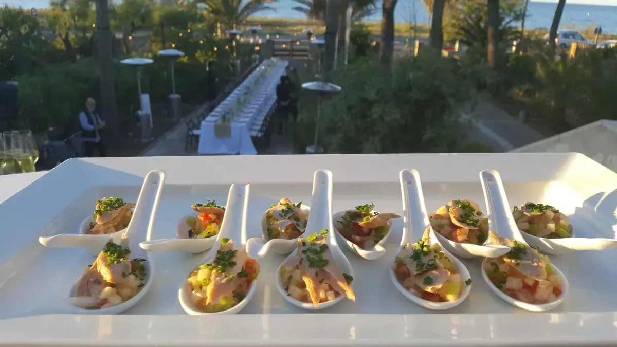 Filas de aperitivos servidos en cucharas blancas sobre una mesa, con un sendero ajardinado y palmeras al fondo.