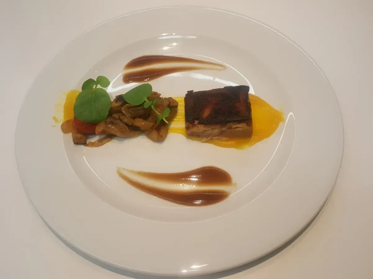 Plato gourmet con carne dorada, salsa amarilla, verduras y espirales decorativas de salsa sobre un plato blanco.