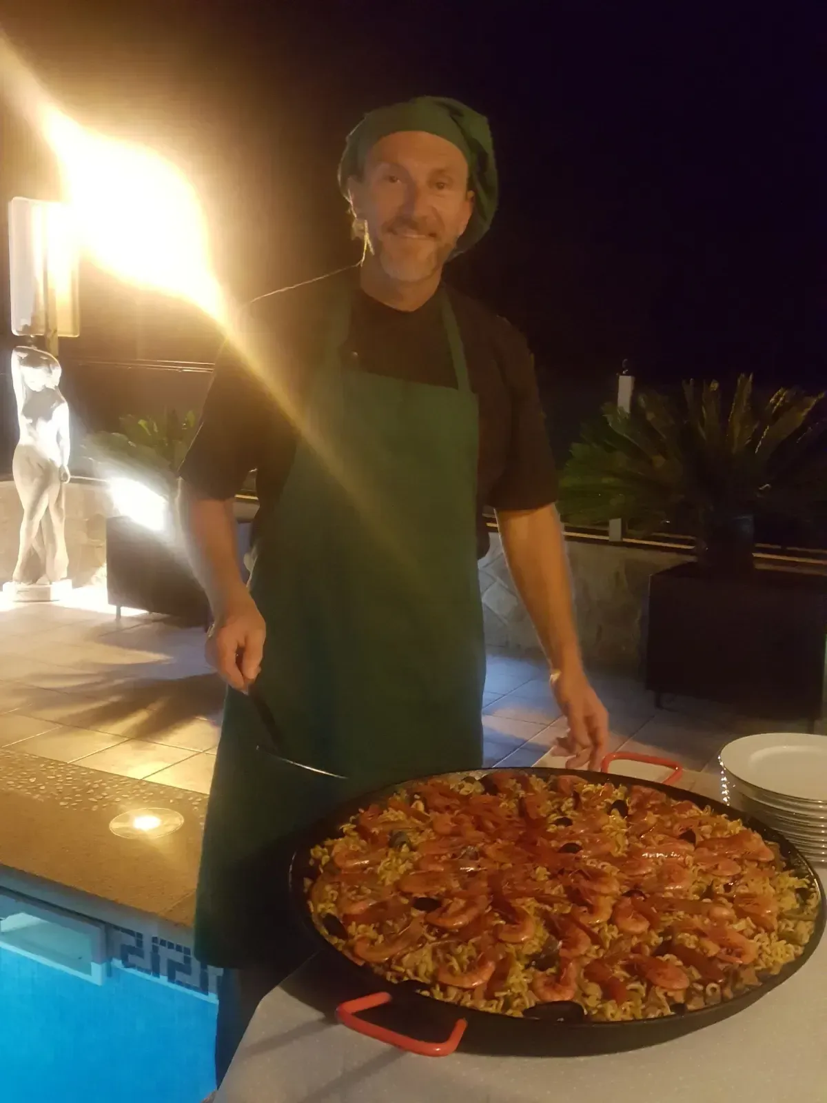 Un chef de pie junto a una piscina iluminada sostiene una gran paella cocinada por la noche.