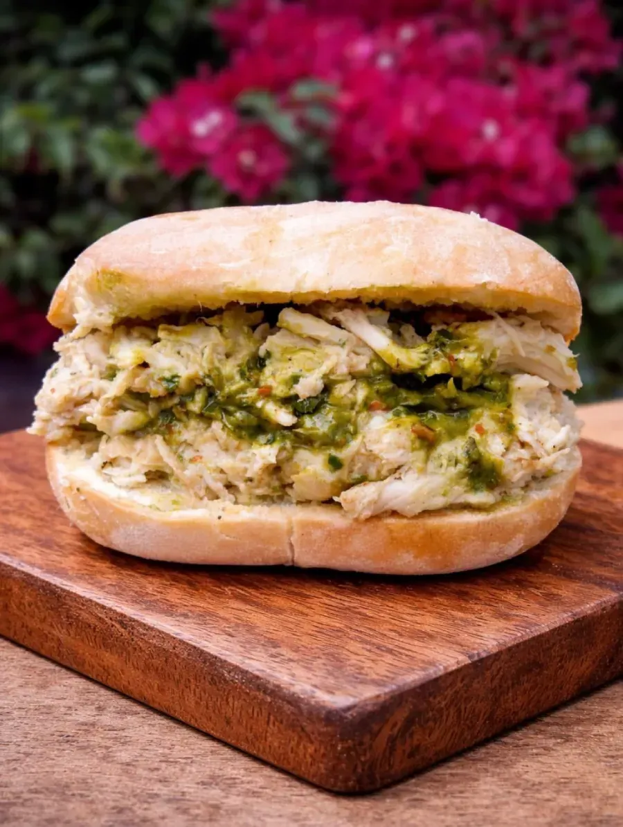Primer plano de un sándwich de pollo sobre una tabla de madera con flores rosas difuminadas al fondo.