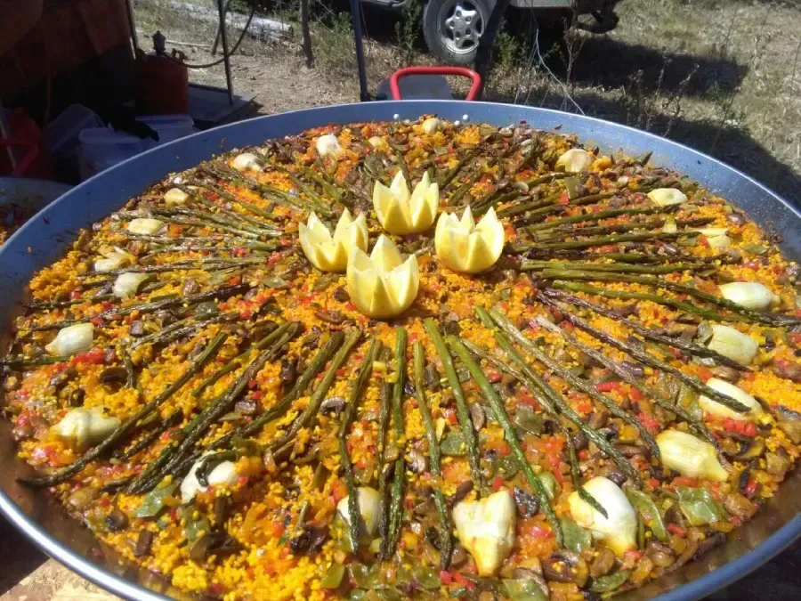 Gran paellera con arroz al azafrán, verduras y rodajas de limón, dispuestas en forma de flor al aire libre.