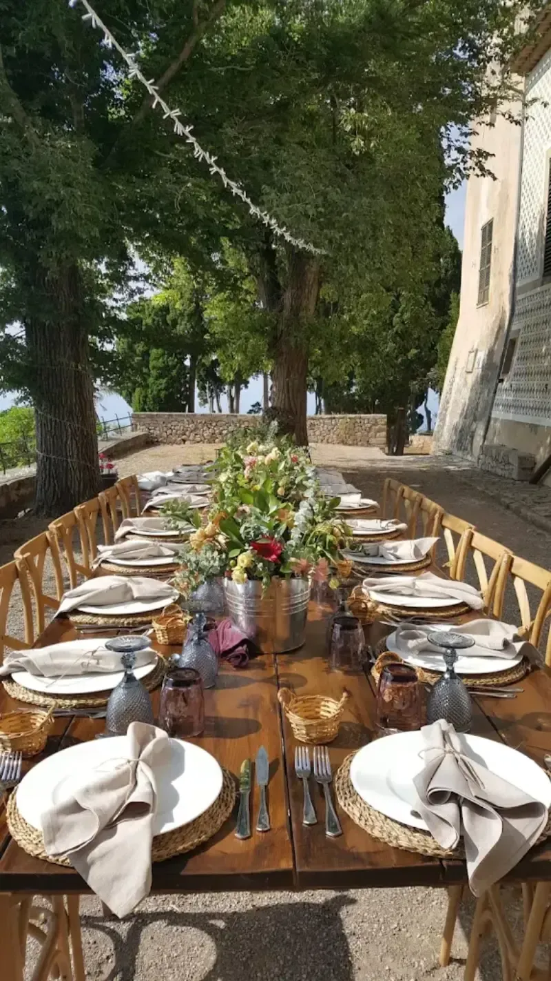 Mesa larga de comedor al aire libre con platos blancos y centro de mesa floral bajo los árboles junto al agua.