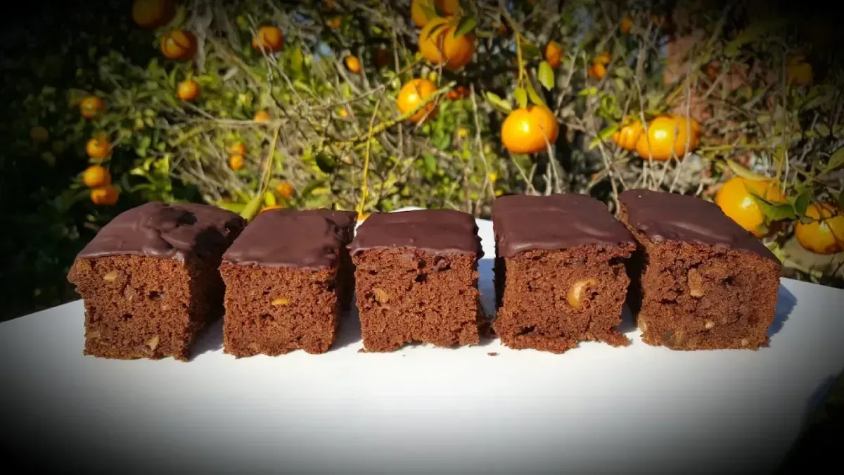 Bizcocho de chocolate en rebanadas con glaseado brillante sobre un plato blanco, con naranjos de fondo.