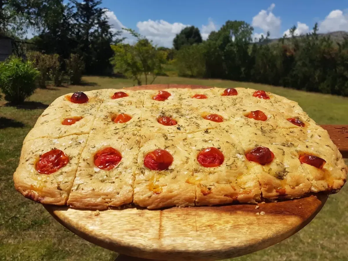 Un pan plano horneado cubierto con tomates cherry reposa sobre una tabla de madera al aire libre.