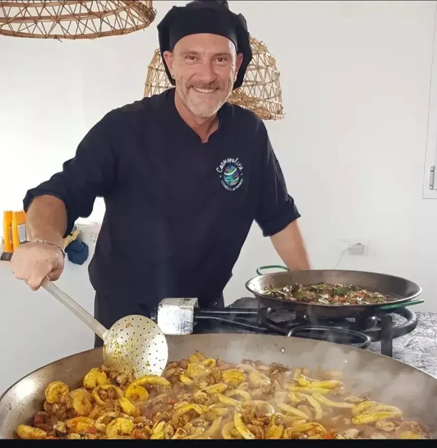 Chef sonriente junto a sartenes humeantes de comida en una cocina luminosa.