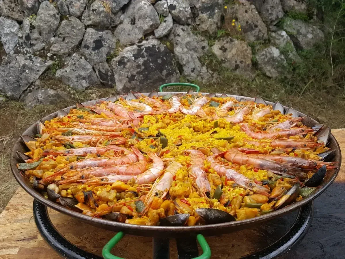 Gran paella de gambas con arroz y limón, cocinada sobre fuego al aire libre contra una pared de piedra.