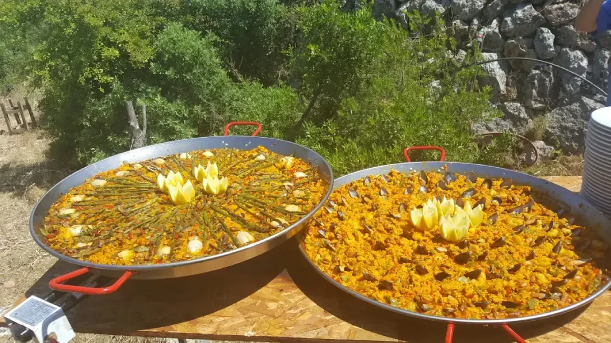 Dos paelleras grandes con arroz amarillo y verduras al aire libre, bajo el sol.