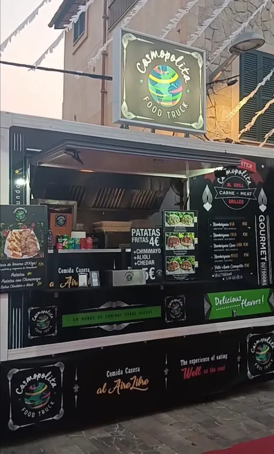 Puesto de comida al aire libre con letreros negros, pizarras de menú iluminadas y platos coloridos en exhibición.