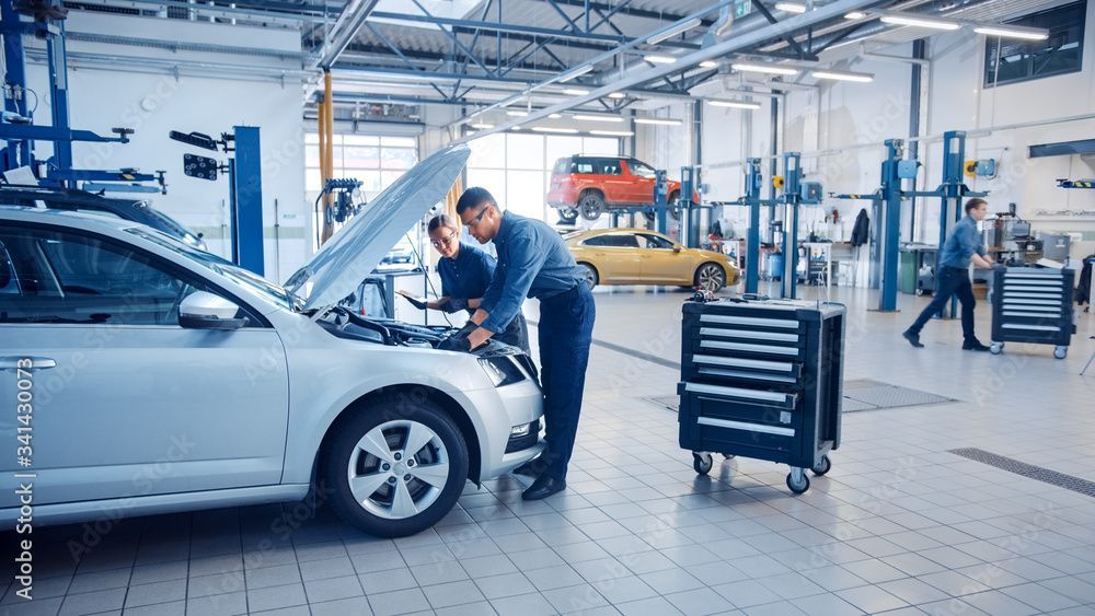 Ein Mechaniker untersucht einen silbernen Wagen in einer hellen Autowerkstatt; im Hintergrund sind Werkzeuge und Fahrzeuge zu sehen.
