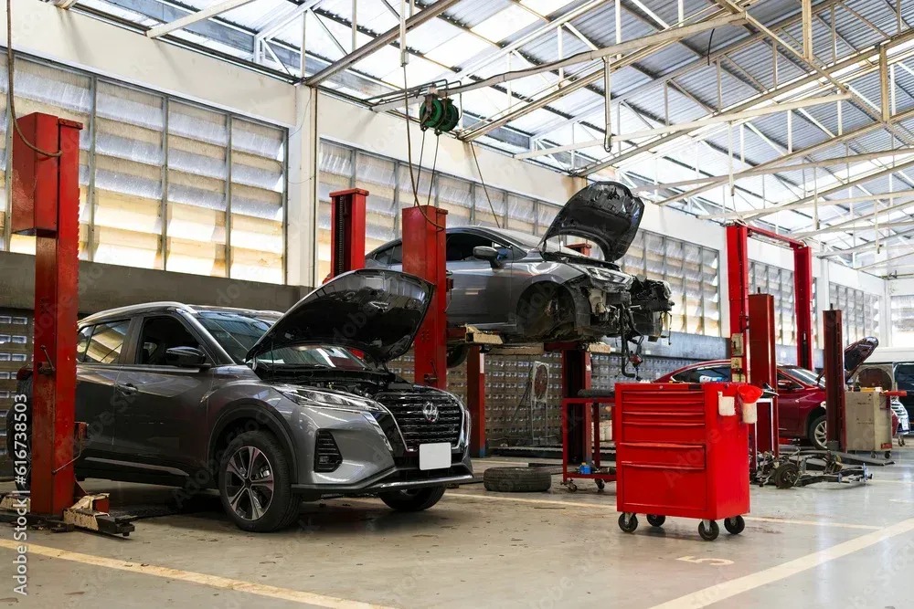 Ein Auto mit geöffneter Motorhaube in einer Autowerkstatt, ein hochgelegter SUV und rote Werkzeugwagen auf einem hellen Werkstattboden
