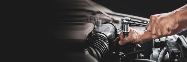 Ein Mechaniker untersucht einen silbernen Wagen in einer hellen Autowerkstatt; im Hintergrund sind Werkzeuge und Fahrzeuge zu sehen.