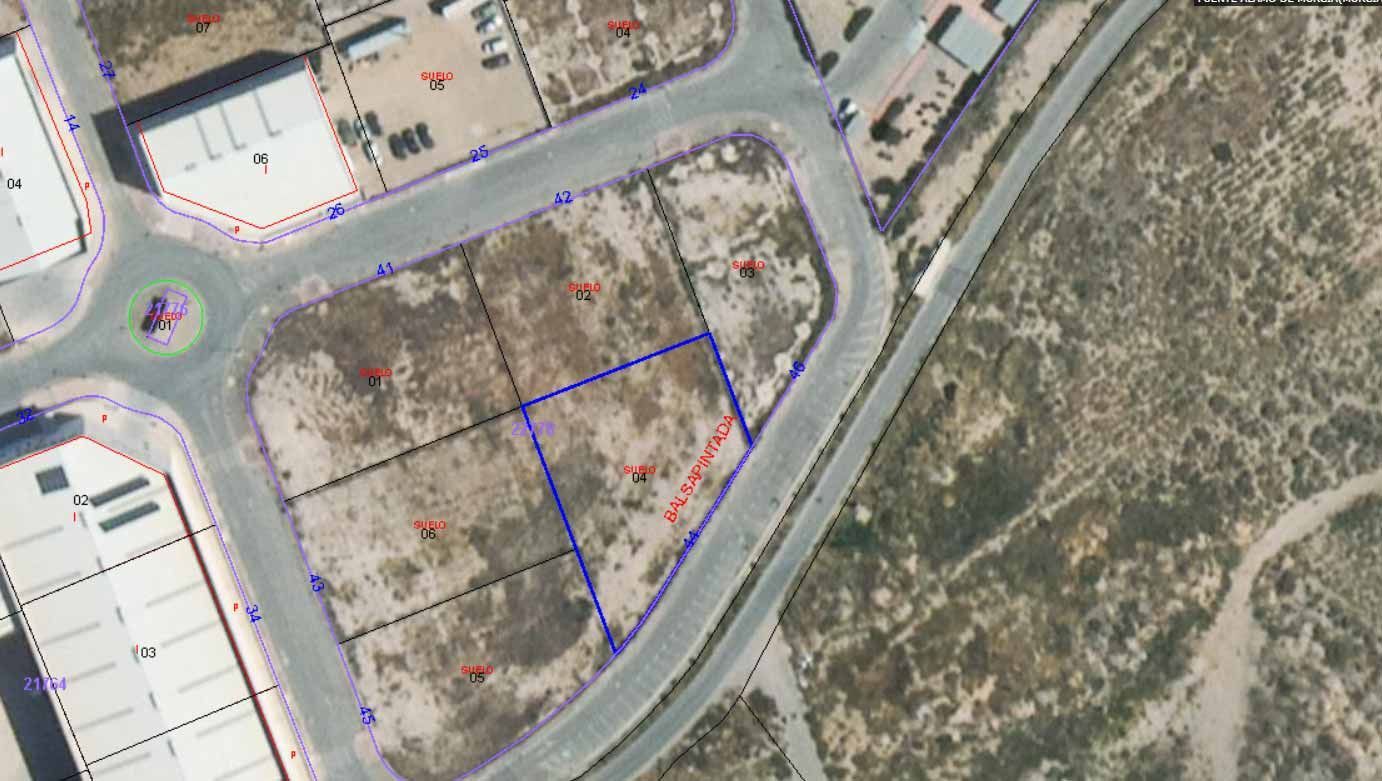 Vista aérea de lotes baldíos delimitados con líneas azules y caminos, algunos con números rojos, cerca de edificios comerciales.