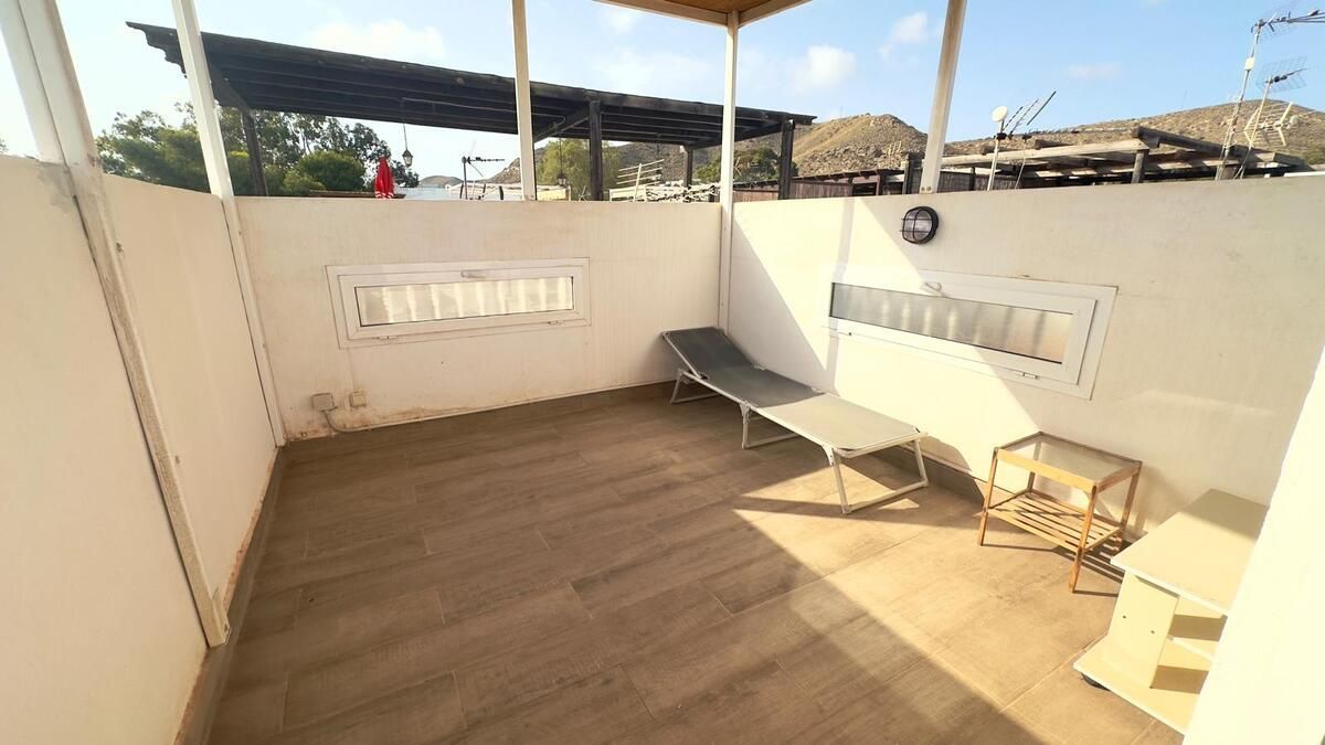 Pequeño espacio al aire libre con un sillón, una mesa pequeña y dos pequeñas ventanas bajo un techo parcial.