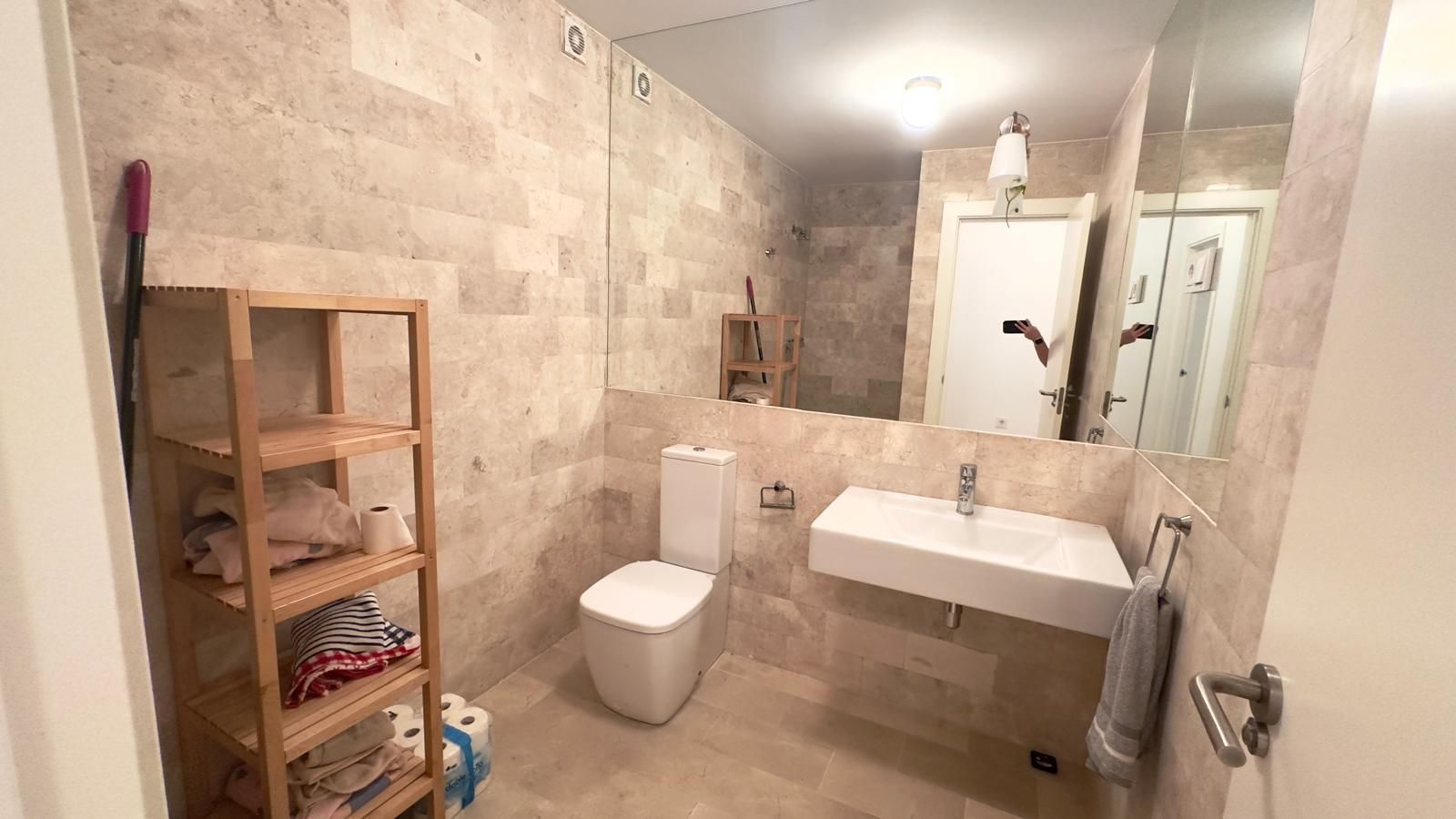 Baño con paredes efecto piedra, lavabo flotante, inodoro y estantería de madera con toallas.