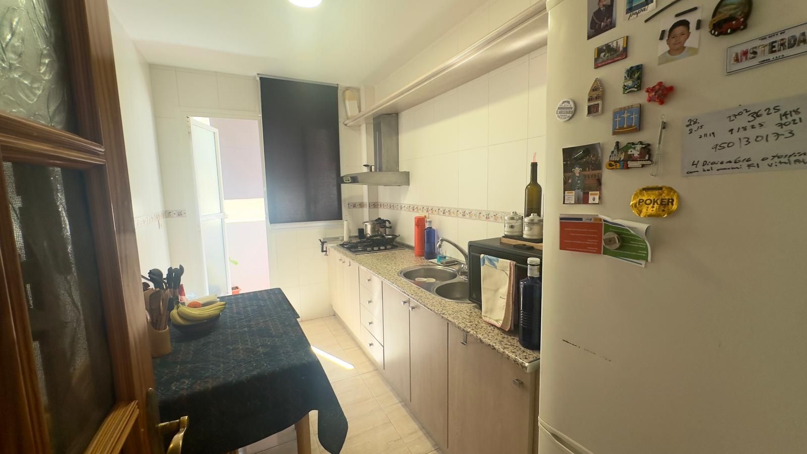 Pequeña cocina con encimera, gabinetes, refrigerador, mesa con frutas y una puerta de entrada.