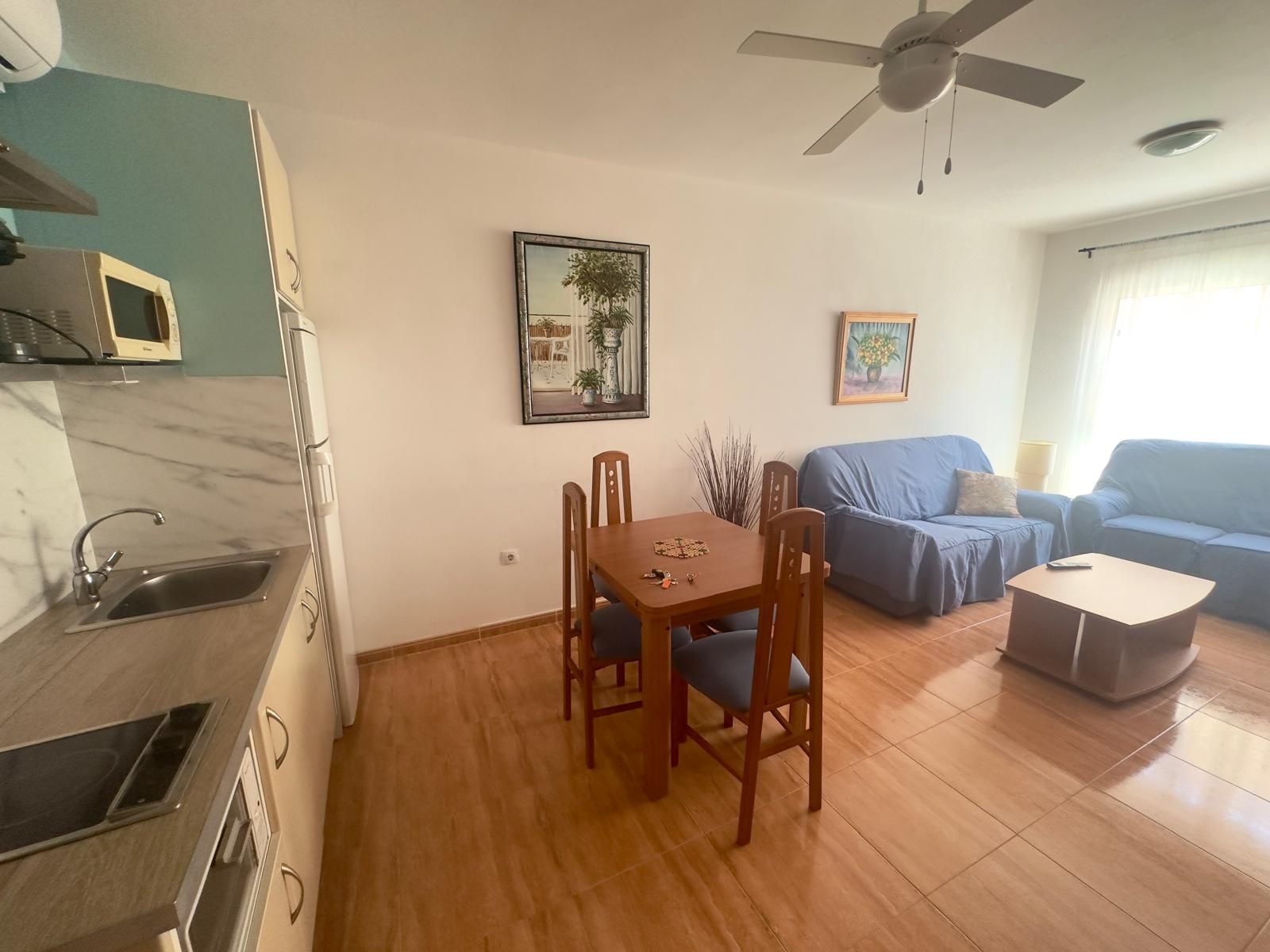Interior de apartamento pequeño con cocina, mesa de comedor, sofás azules, obras de arte y ventilador de techo.
