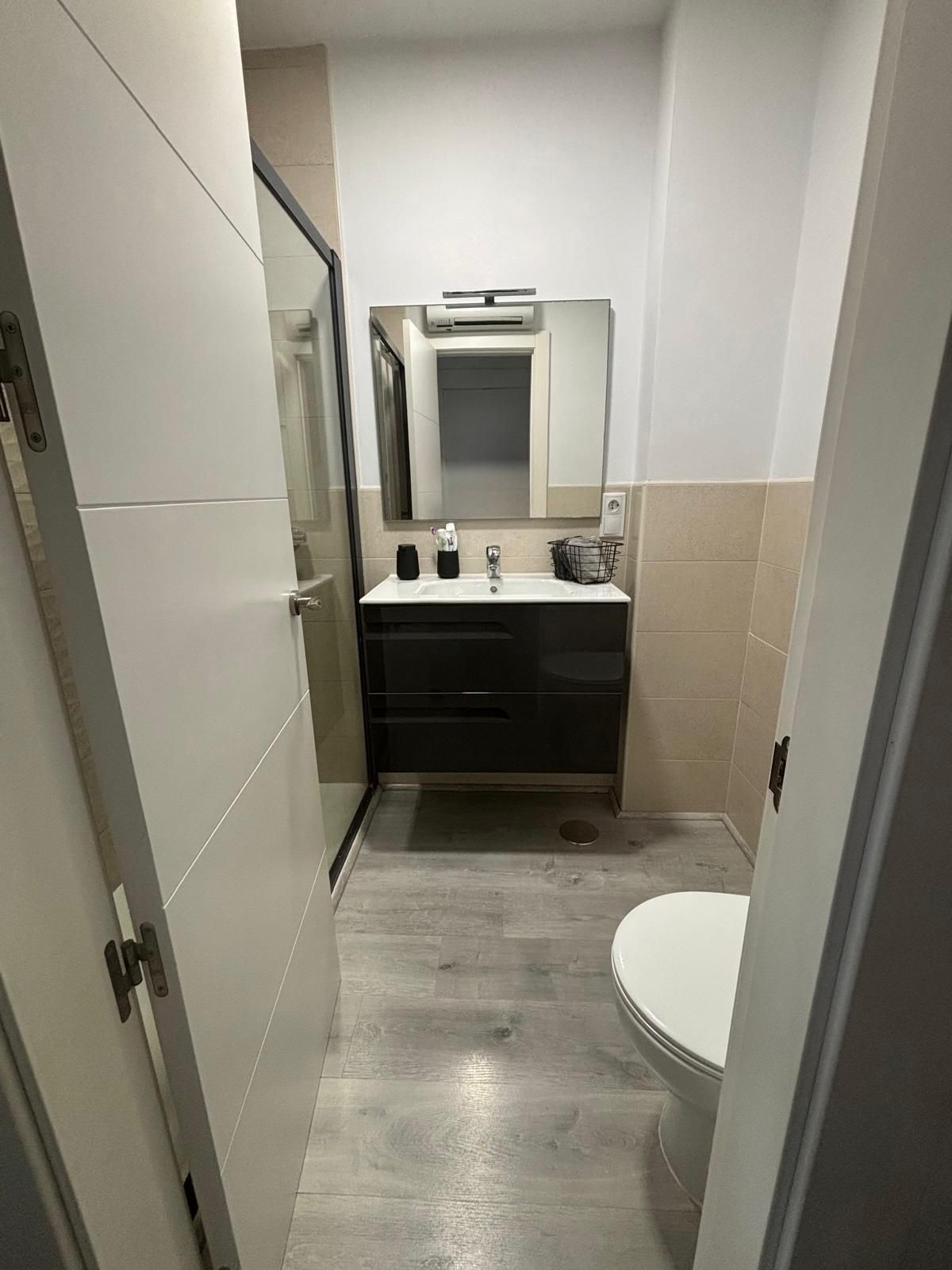 Baño estrecho con inodoro blanco, lavabo oscuro y ducha de cristal. Suelo gris, azulejos beige y paredes blancas.