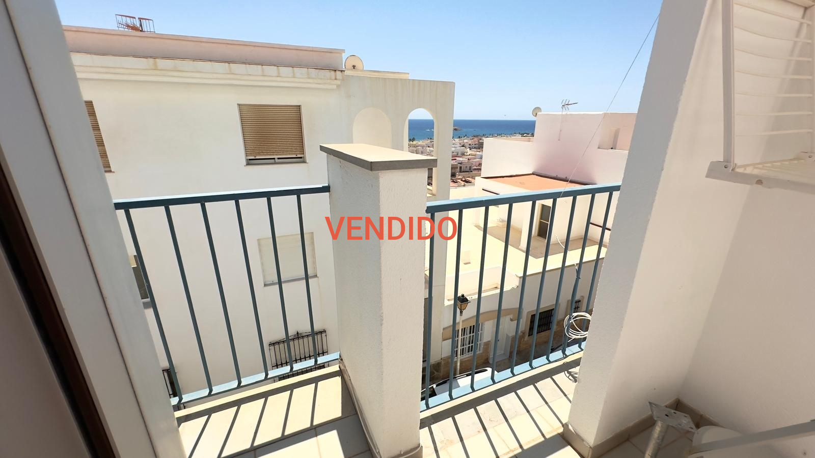 Balcón con vista al paisaje urbano y al océano, con barandilla azul, edificios blancos y cielo despejado.