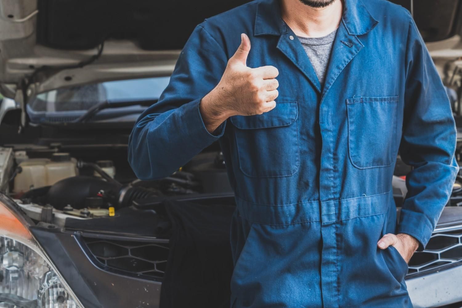 Un mecánico con mono azul está de pie delante de un coche con el capó abierto, haciendo un gesto de aprobación con el pulgar.