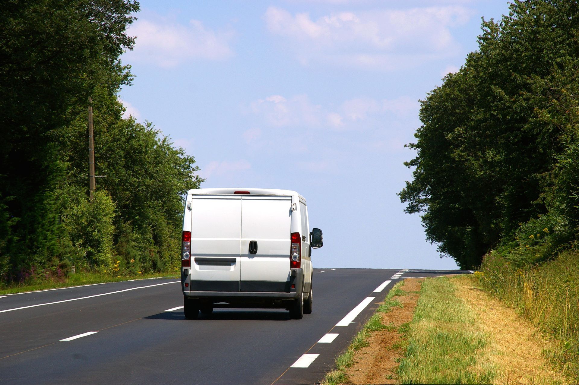 Une camionnette blanche circule sur une route goudronnée, bordée d'arbres, par une journée ensoleillée.
