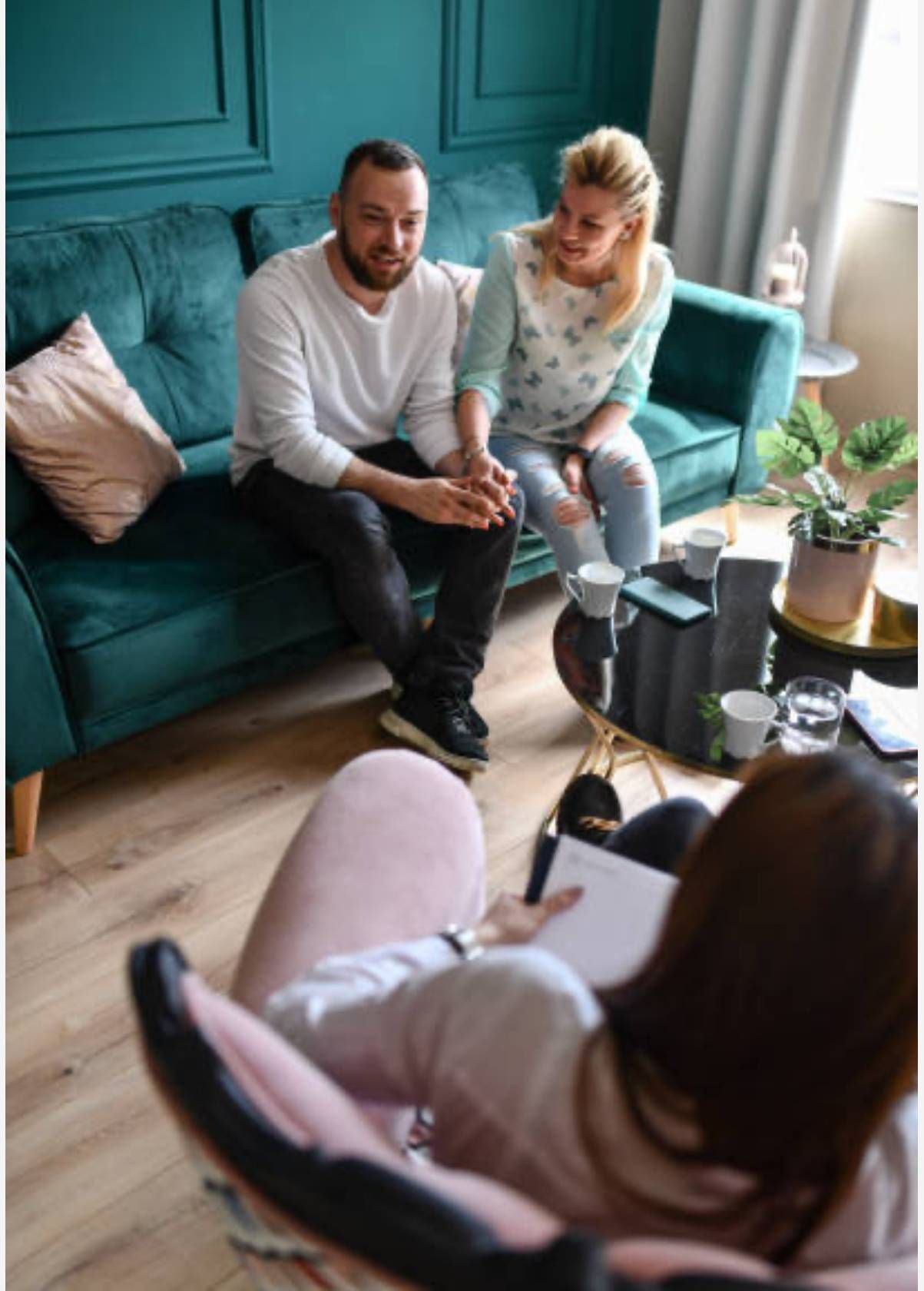Pareja en un sofá verde azulado hablando con una persona en una silla rosa, posiblemente un terapeuta.