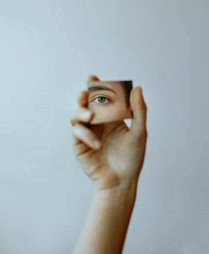 Mano sosteniendo un pequeño espejo que refleja un ojo azul sobre un fondo blanco.