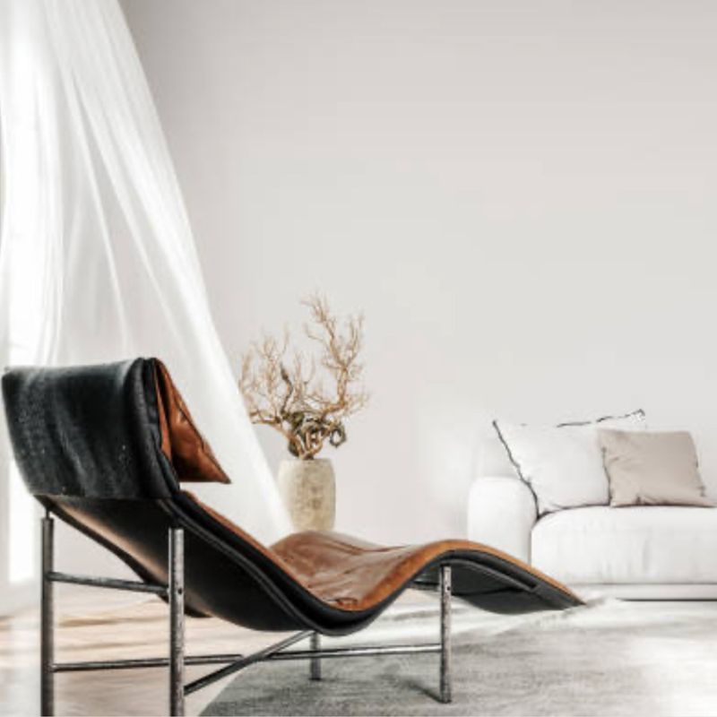 Chaise longue de cuero en una habitación luminosa con un sofá blanco y un jarrón con flores secas.