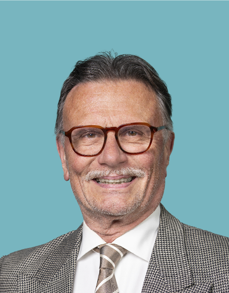 Mann mit Brille, lächelnd, in gemustertem Blazer und Krawatte, vor blaugrünem Hintergrund.