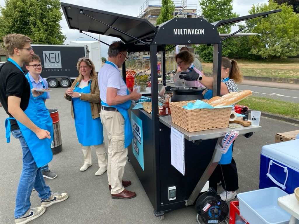 Menschen an einem Imbissstand. Einige tragen blaue Schürzen und bereiten im Freien Essen zu und kaufen es ein.