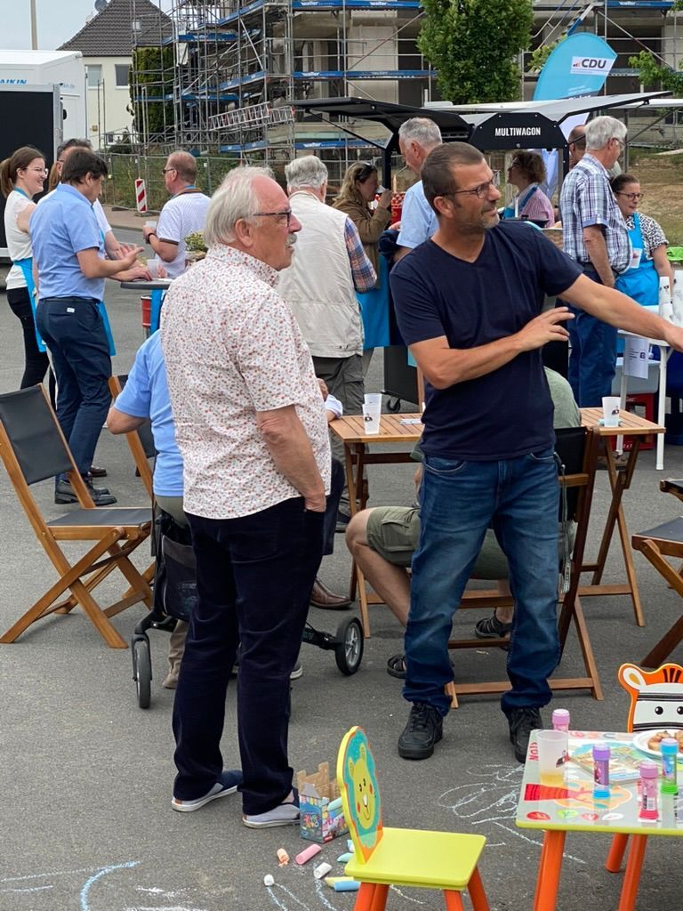 Zwei Männer unterhalten sich bei einer Veranstaltung im Freien mit einer Menschenmenge. Einer zeigt nach rechts, während der andere zuhört.