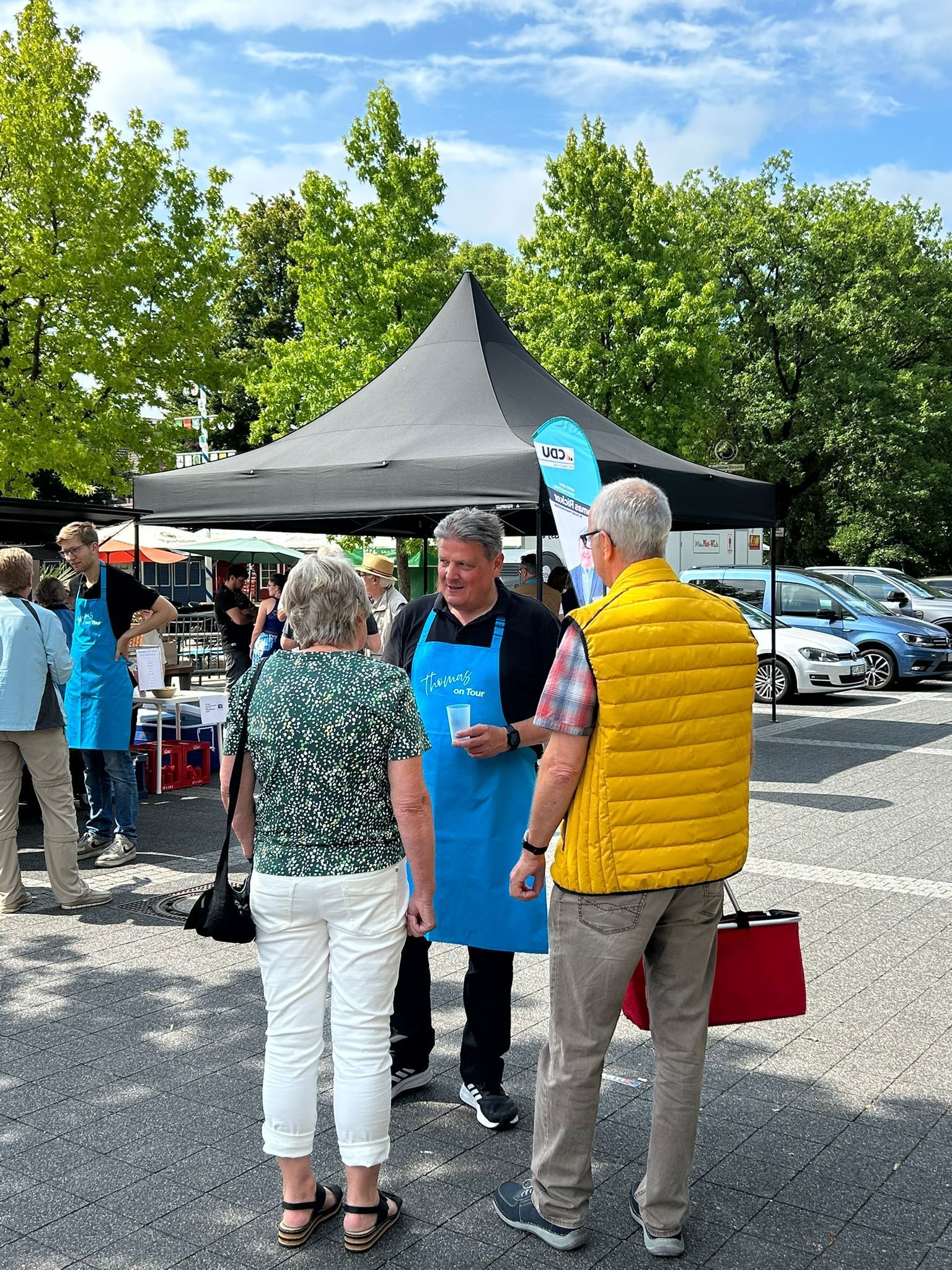 Ein Mann in einer blauen Schürze spricht mit zwei Personen auf einem Markt im Freien unter einem schwarzen Baldachin; sonniger Tag.
