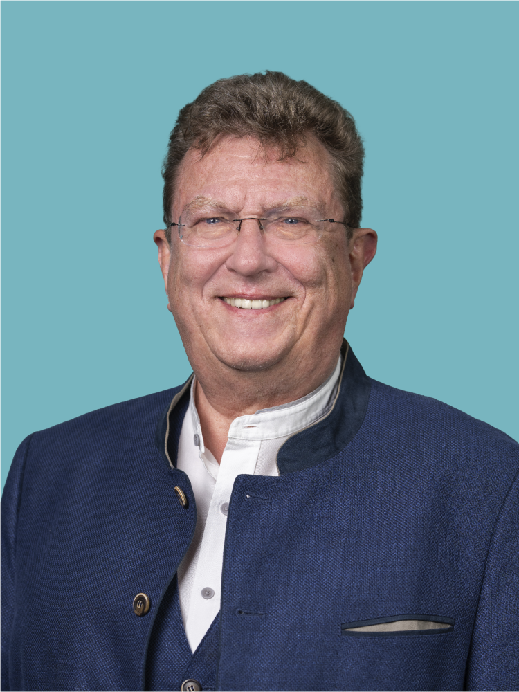 Lächelnder älterer Mann in dunklem Blazer und blauem Hemd steht im Freien vor einem unscharfen Bücherregal im Hintergrund.