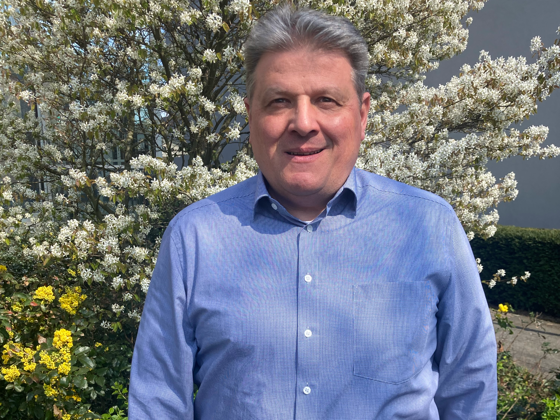 Ein Mann in einem blauen Hemd steht vor einem Baum mit weißen Blüten.