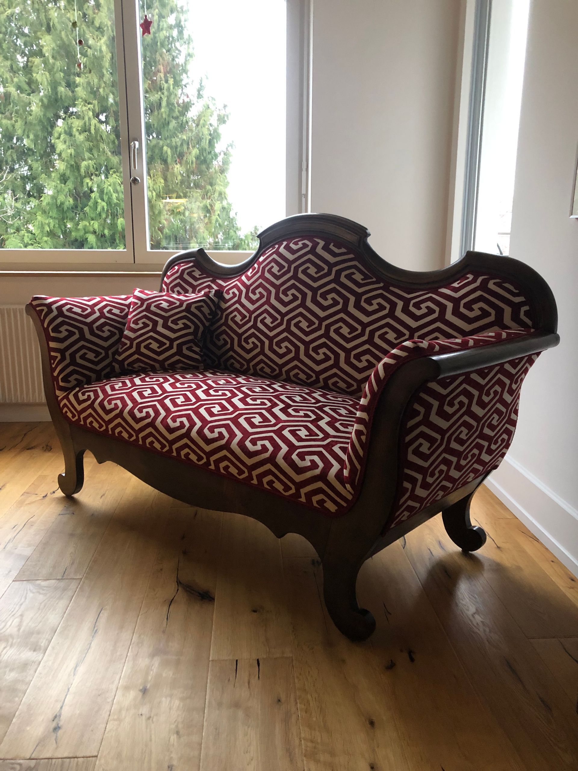 Eine rot-schwarze Chaiselongue steht auf einem Holzboden vor einem Fenster.