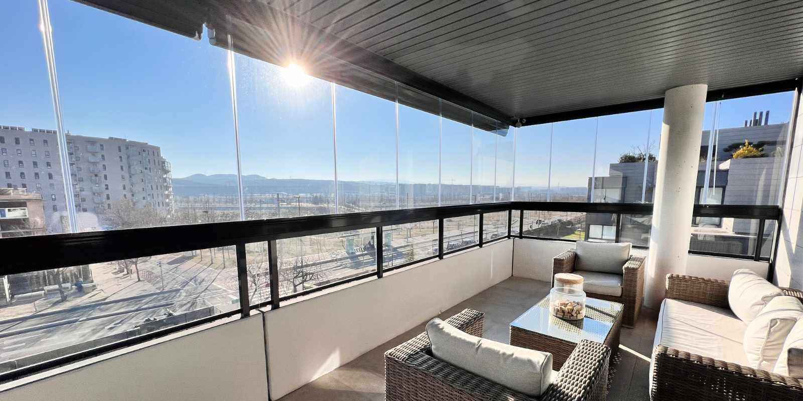 Balcón con ventanales, vista a la ciudad, muebles de mimbre, día soleado.