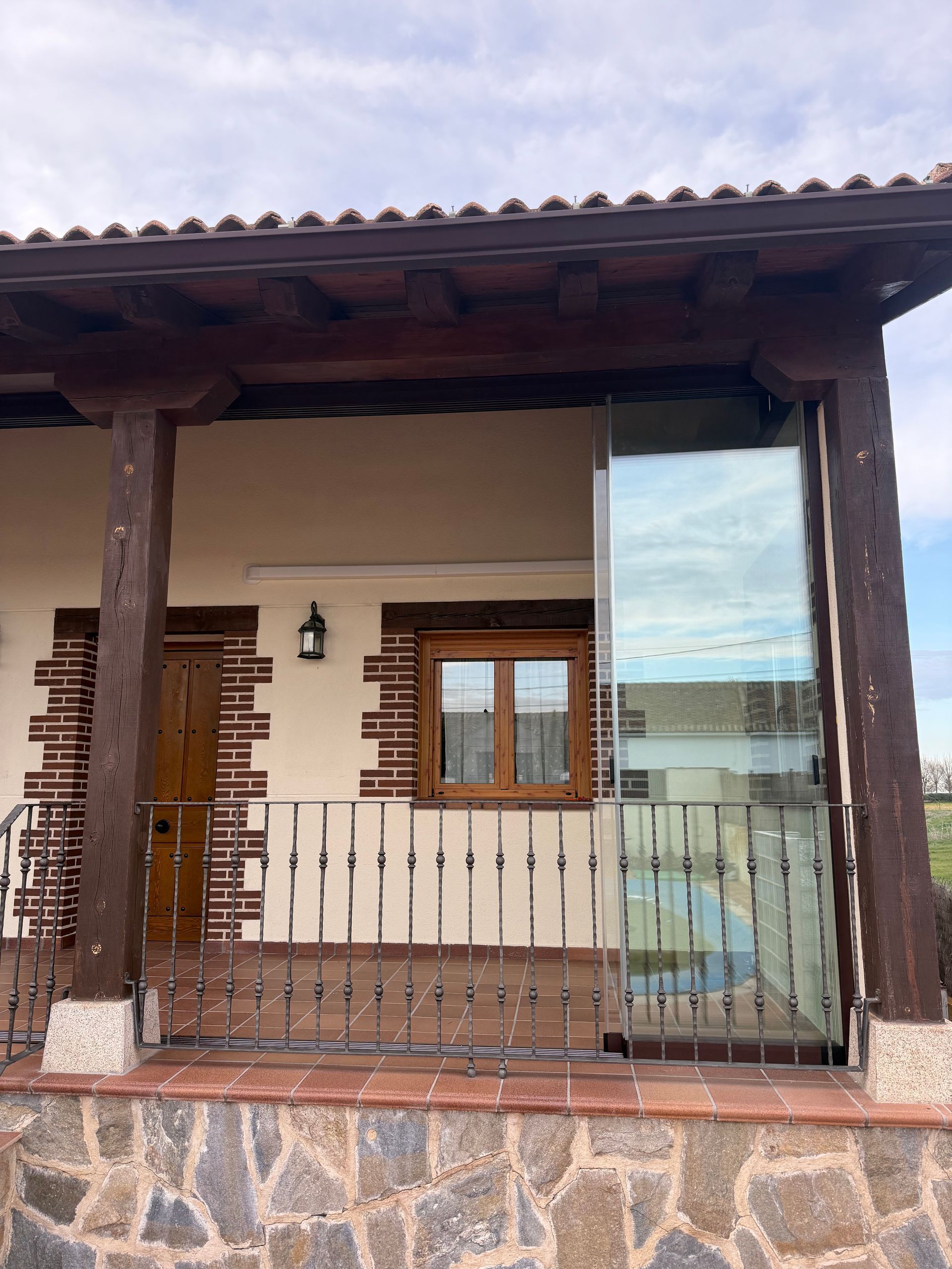 Terraza acristalada con paredes de cristal, sofá gris, mesa redonda y suelo de madera, con vistas a un patio verde.