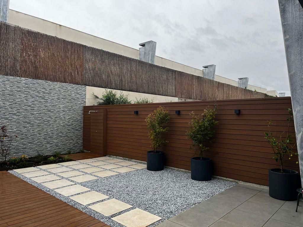 Patio con cerca de madera, muro de piedra, grava, adoquines y árboles en macetas.