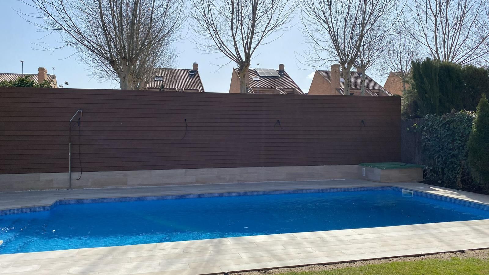 Una piscina rectangular de agua azul frente a una valla de madera y casas bajo un cielo azul.