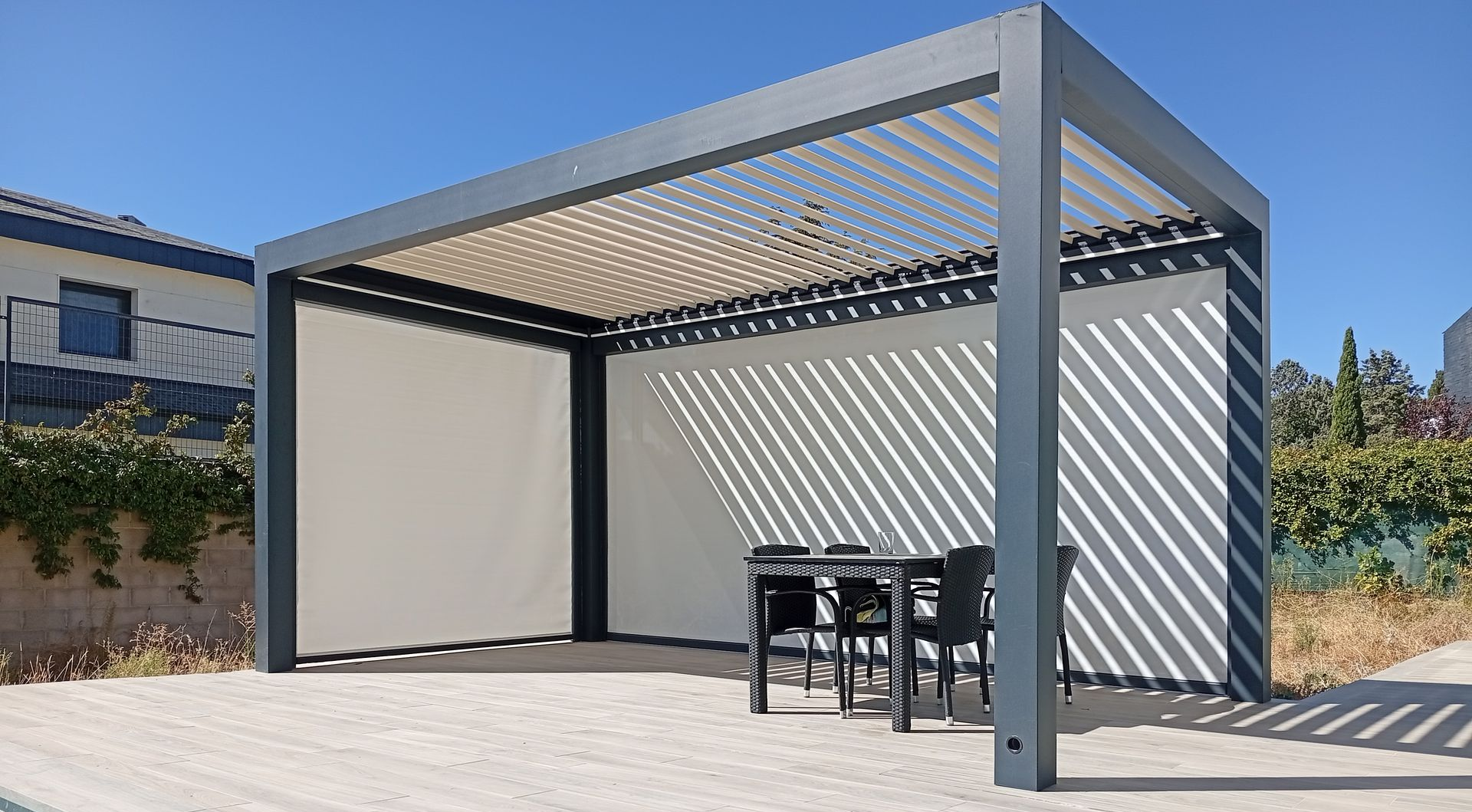 Pérgola gris sobre una zona pavimentada con césped artificial. Día soleado.